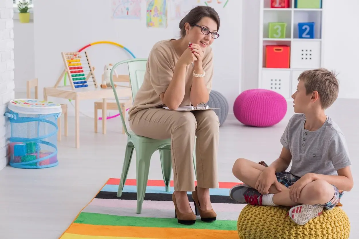 A young boy sitting on a pouf talking to a school psychologist